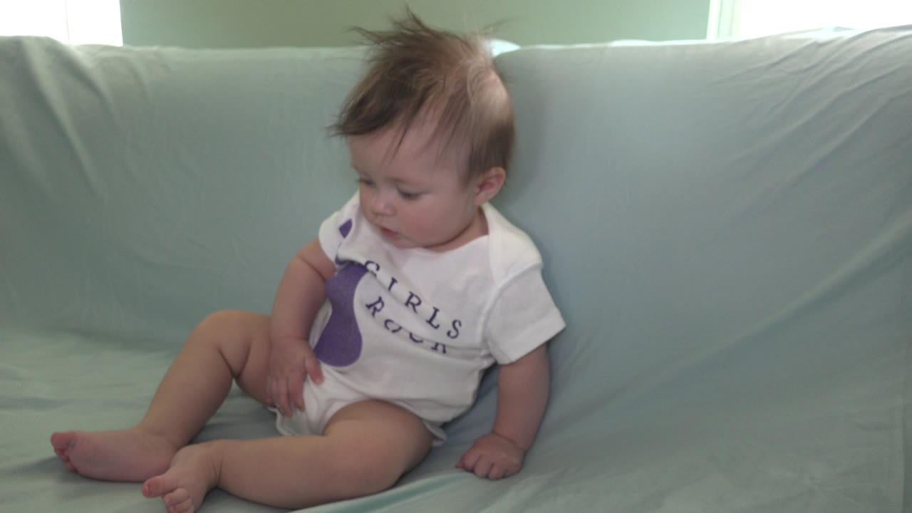 A baby sits near the left side of a pale blue couch in Westminster, Colorado, looking down with one hand near the leg and soft window light behind the cushions.