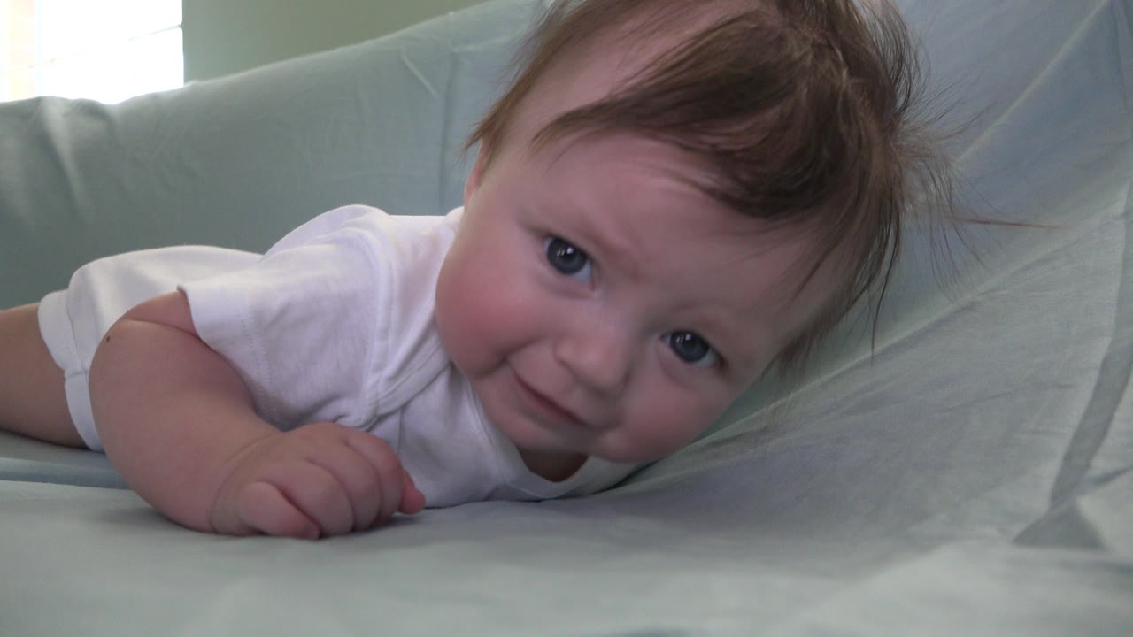 Newborn baby lying face down on pale bedding and looking toward the camera.