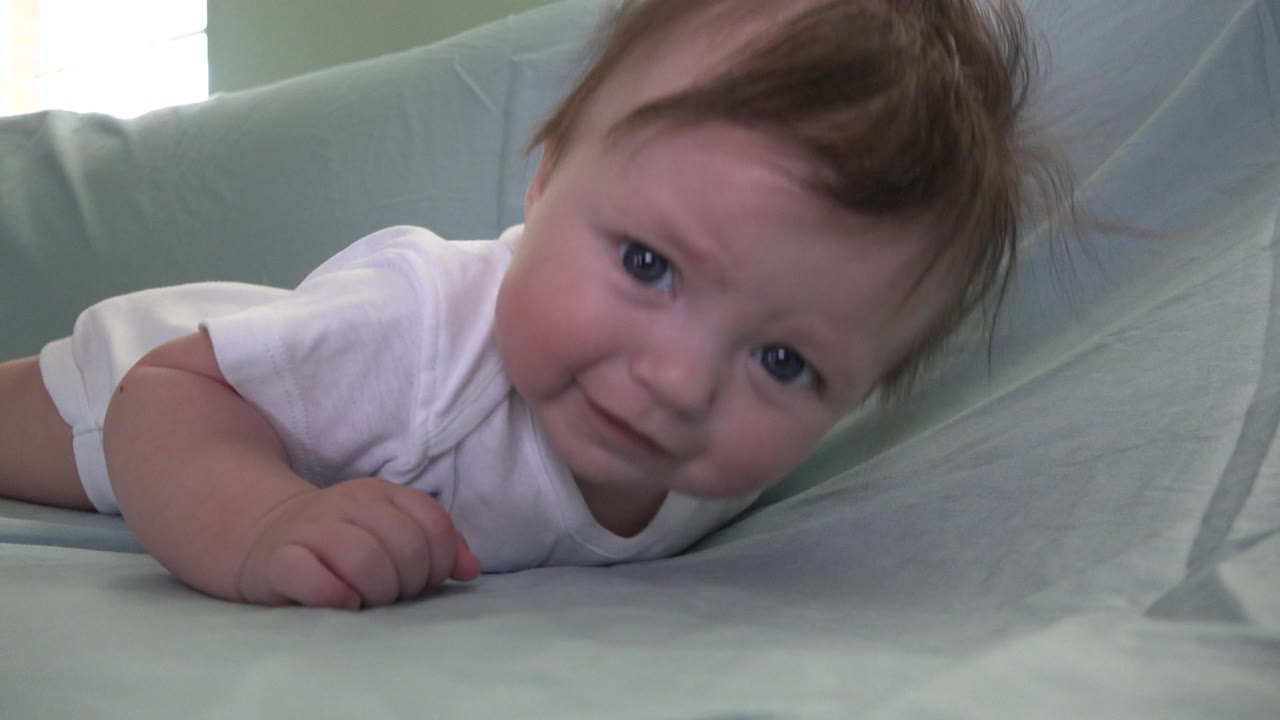 The baby remains belly-down on pale bedding in Westminster, Colorado, with eyes open, cheeks visible, and one hand pressed into the sheet.