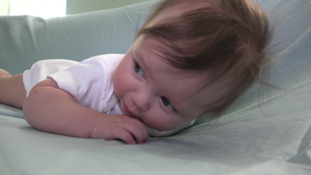 Baby lying on a blue couch with fingers near the mouth.