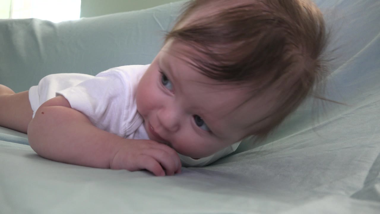 At 0:01 in Westminster, Colorado, the baby remains on the blue couch, looking off to the side while fingers press against the mouth.