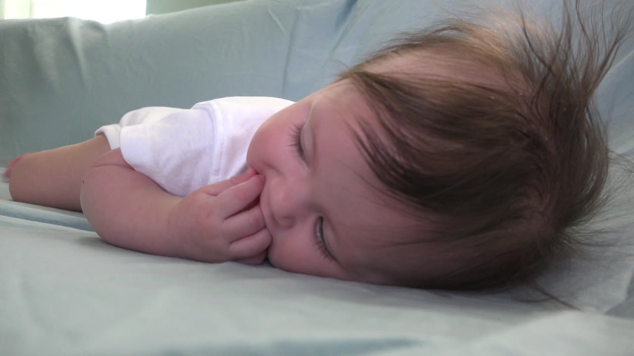 At 0:00 in Westminster, Colorado, an infant girl lies sideways on pale blue bedding with her fingers near her mouth and her hair spread toward the top of frame.