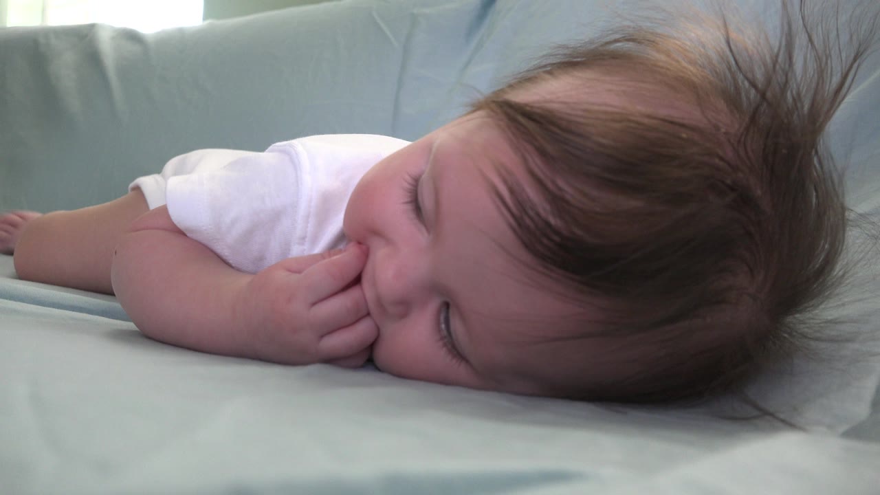 At 0:01 in Westminster, Colorado, the infant remains on her side on the pale blue surface, with one eye partly visible and her hand pressed near her lips.