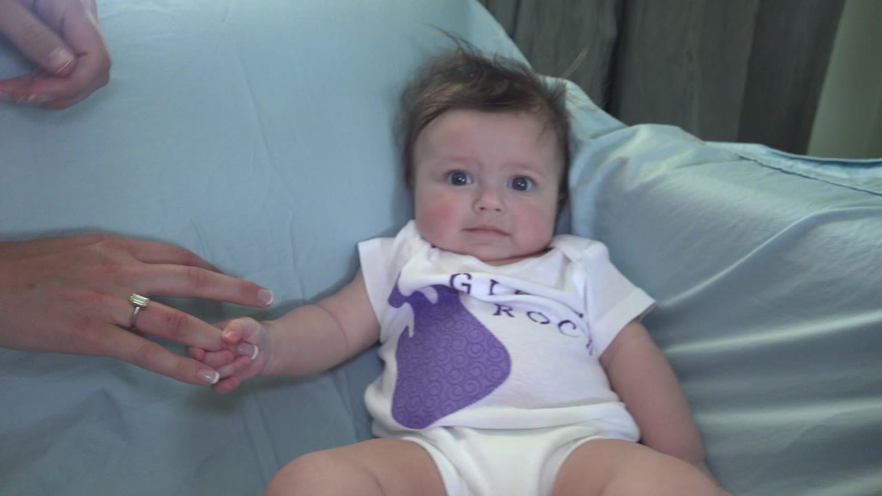 At 0:00 in Westminster, Colorado, a baby in a white onesie lies against pale blue couch cushions while an adult hand holds the baby's hand.