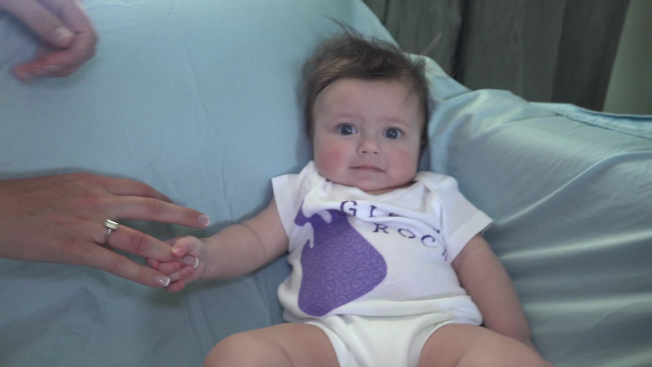 At 0:01 in Westminster, Colorado, the baby looks toward the camera from the couch as an adult hand rests near the baby's left hand.