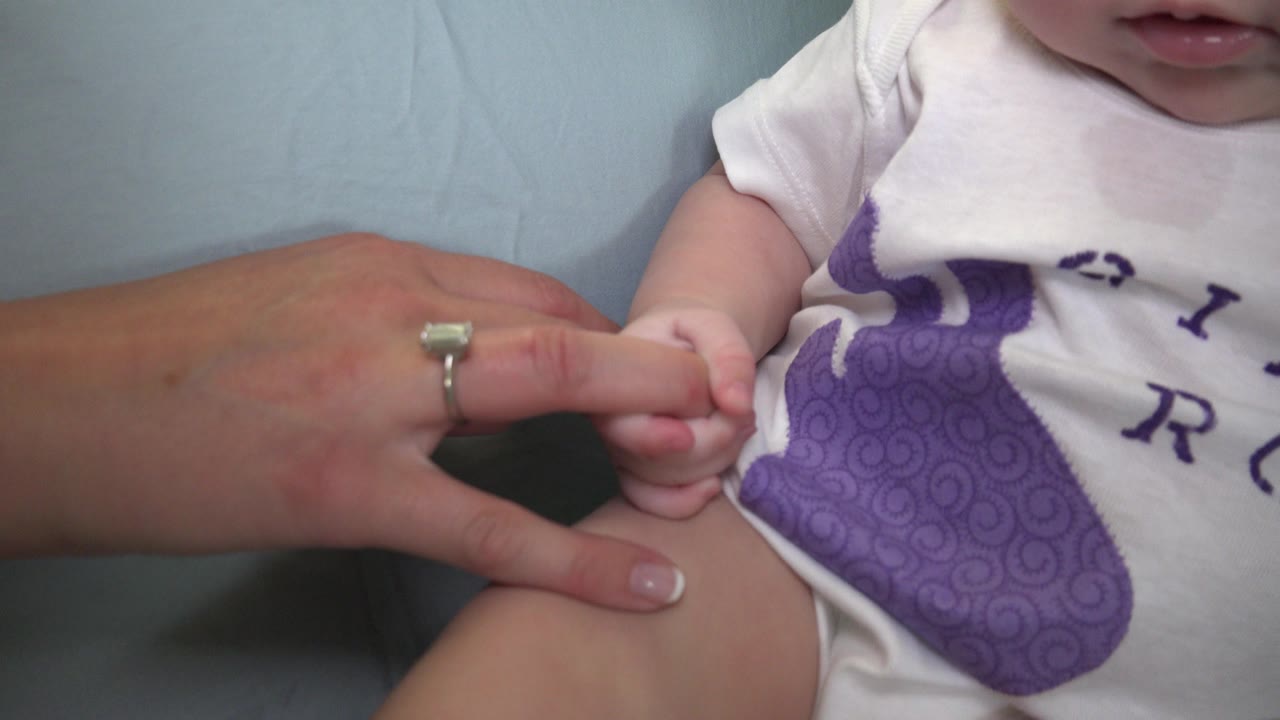 Close frame from Westminster, Colorado shows a baby in a white onesie gripping an adult woman's finger beside the baby's bent leg on pale blue fabric.