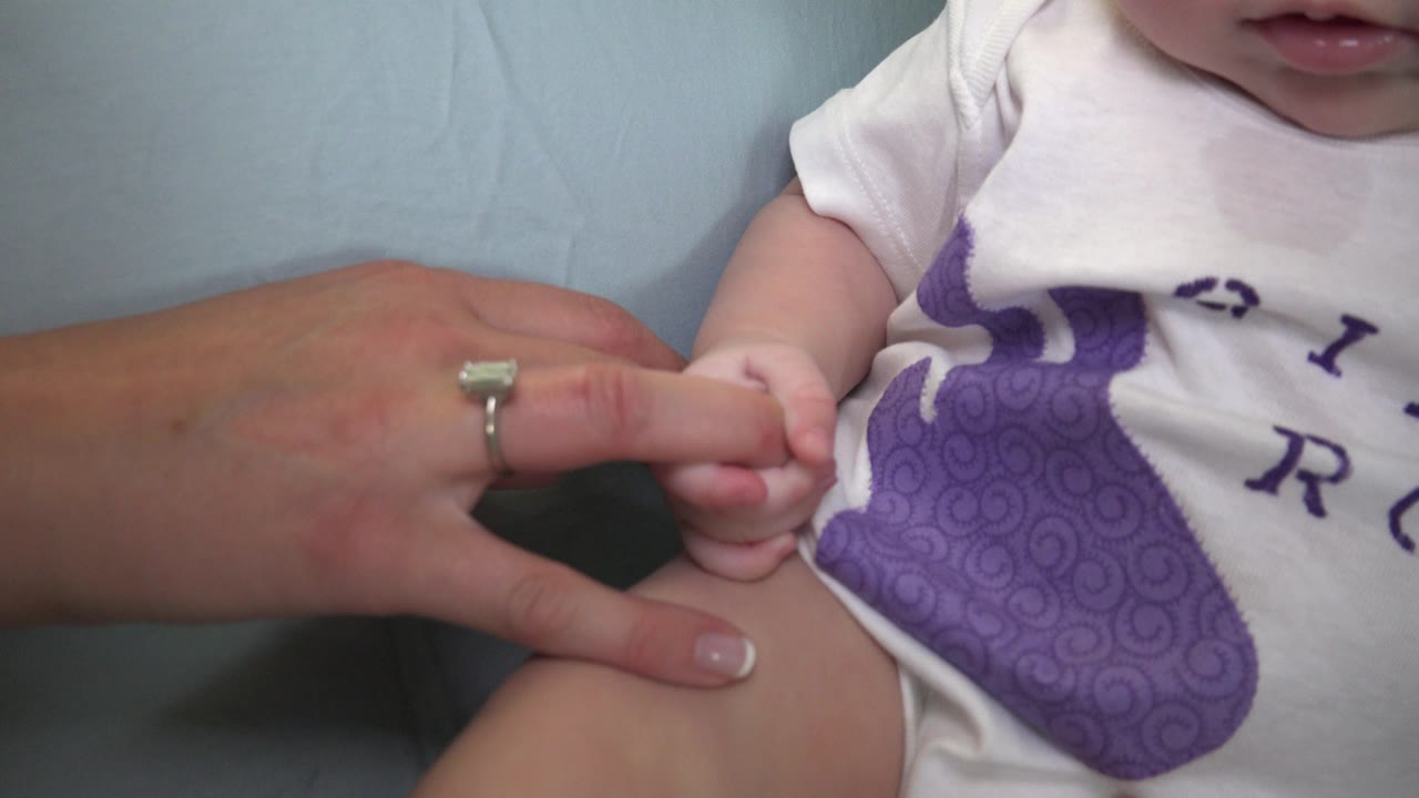 The baby remains cropped at the mouth and torso while the adult woman's hand rests across the foreground near the infant's fingers and white onesie.