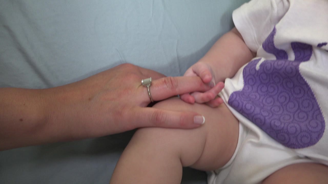 At 0:00 in Westminster, Colorado, an adult hand holds a baby's fingers near the baby's bent leg and white onesie with a purple graphic.