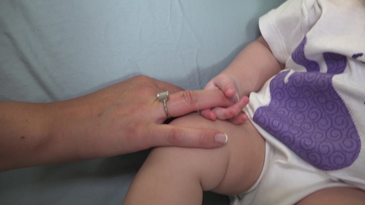 At 0:01 in Westminster, Colorado, the adult hand remains on the baby's fingers, with the white onesie and purple chest graphic centered in the tight frame.