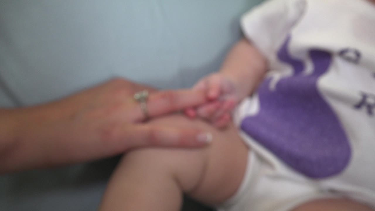 Soft-focus close-up of an adult hand and baby's fingers near a purple graphic on a onesie.