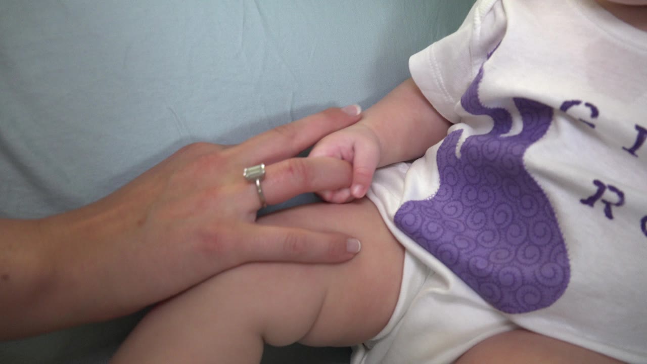 The adult hand remains on the baby's bent leg in this Westminster, Colorado close-up, with the white onesie and purple graphic filling the right side of frame.