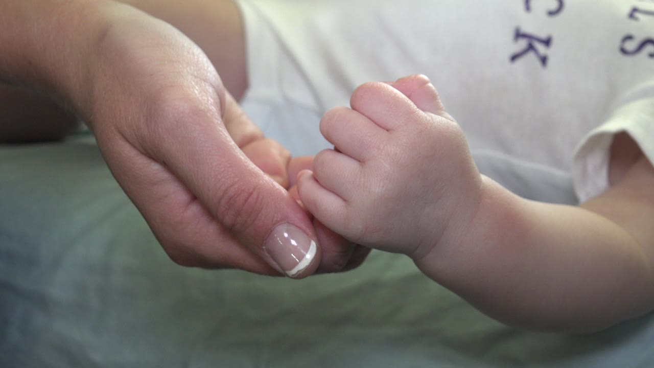 At the start of the Westminster, Colorado clip, an adult hand lightly holds a baby's foot against green bedding with a blurred white shirt behind it.