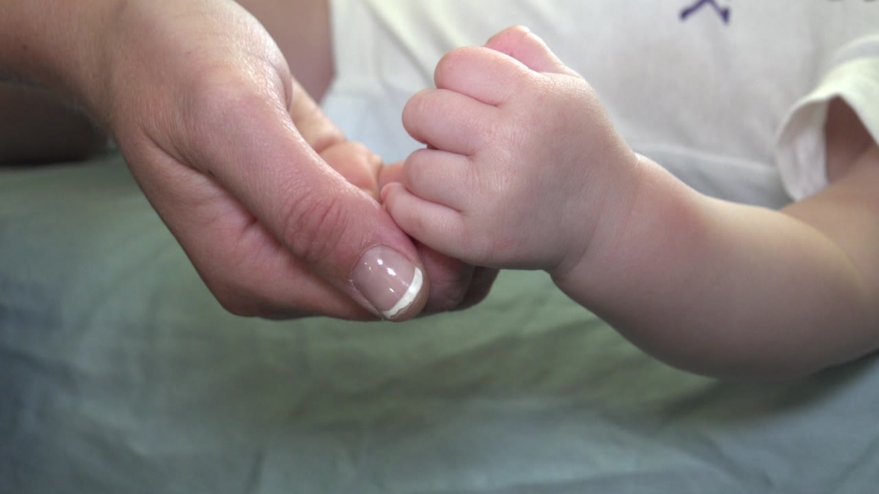 At 0:01 in Westminster, Colorado, the baby's curled fingers remain wrapped around an adult finger, with a white shirt visible behind the hands.