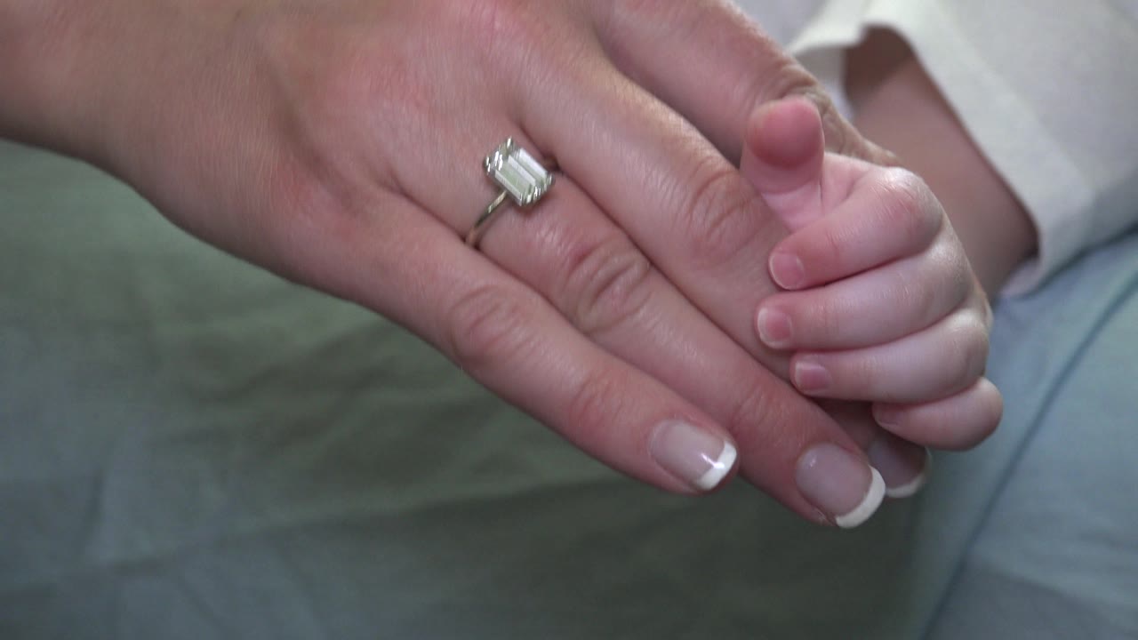 The ringed adult hand remains close to the infant's hand, with the diamond catching soft light and the white sleeve and blue fabric blurred behind it in Westminster, Colorado.