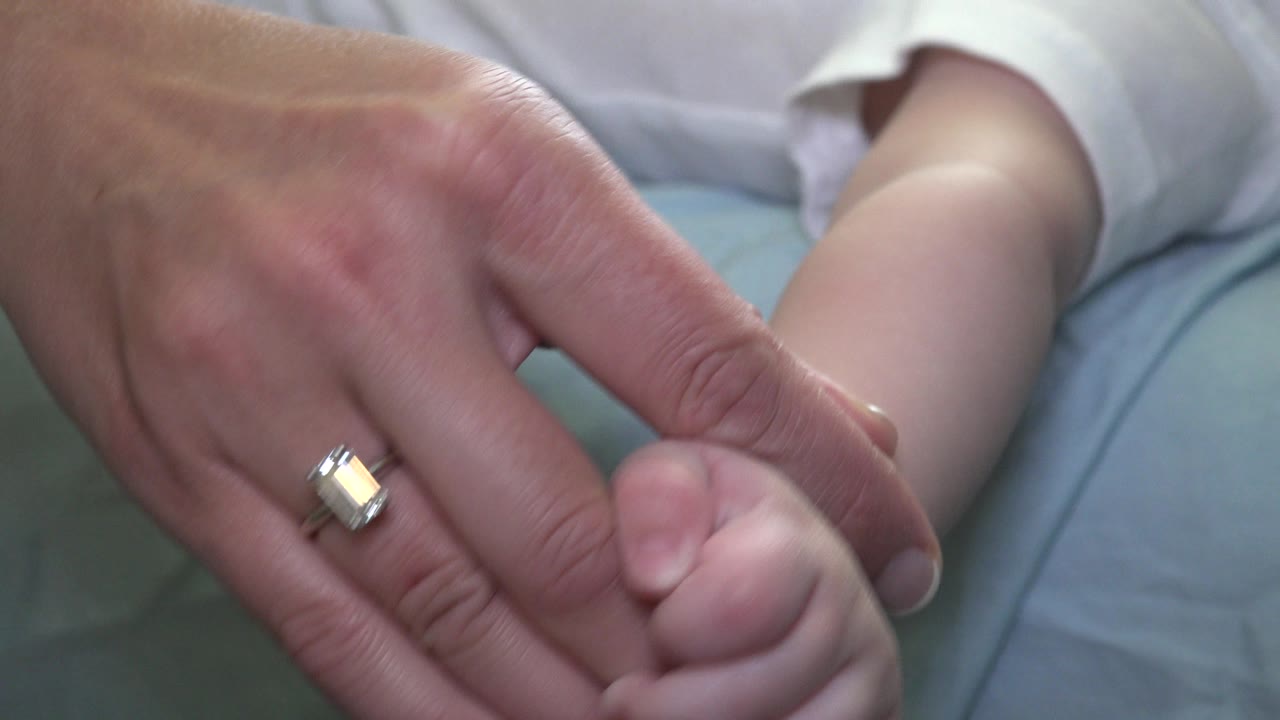 In Westminster, Colorado, the ringed adult hand remains close to the child's curled hand, with soft bedding and a white sleeve behind them.