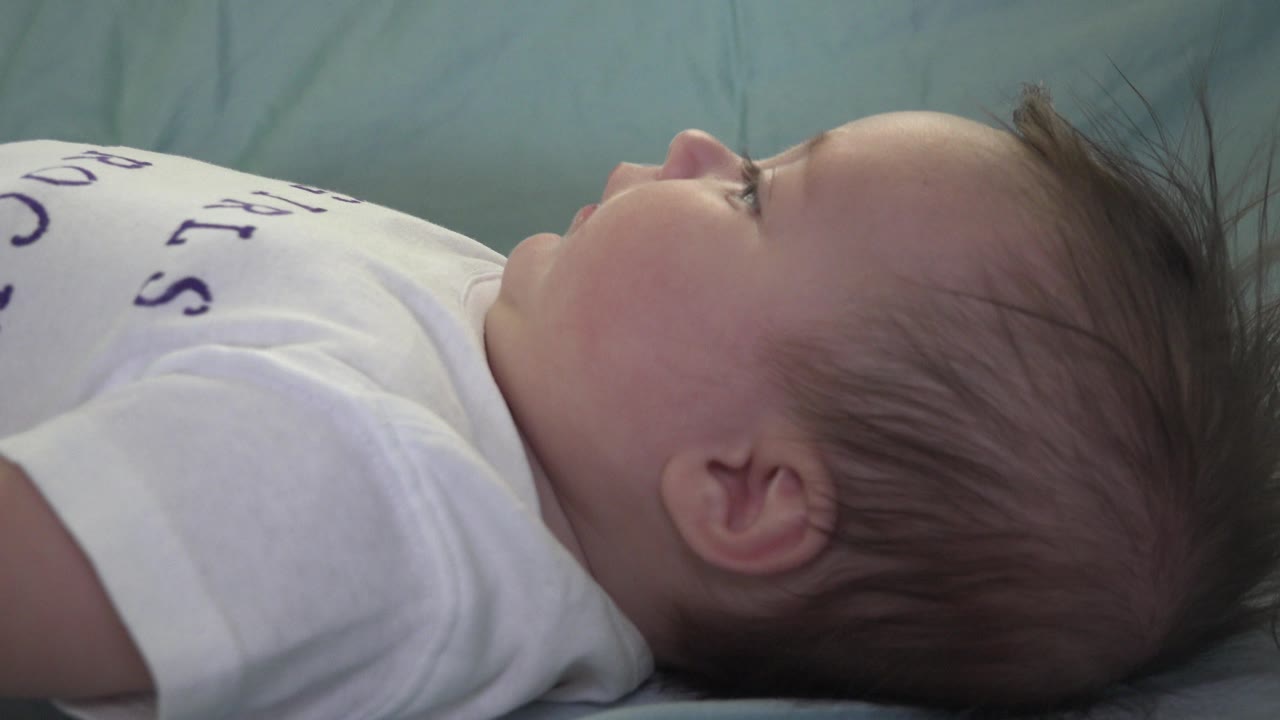 At 0:01, the baby's face remains turned upward in profile on the bed, with the ear and wisps of hair more central in the close frame.