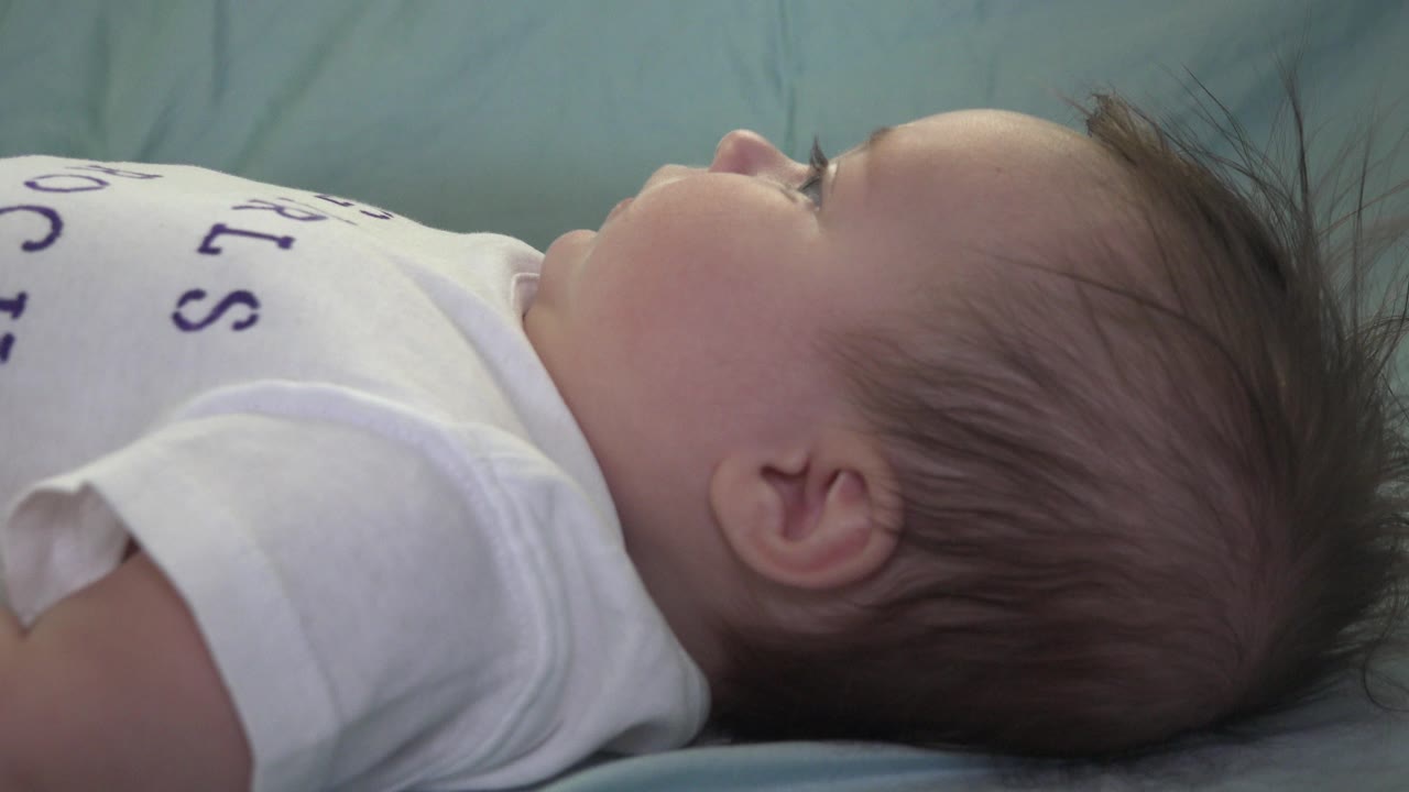 At 0:01 in Westminster, Colorado, the infant stays reclined on the blue bedding, with the cheek, ear, hair, and white shirt filling the tight frame.