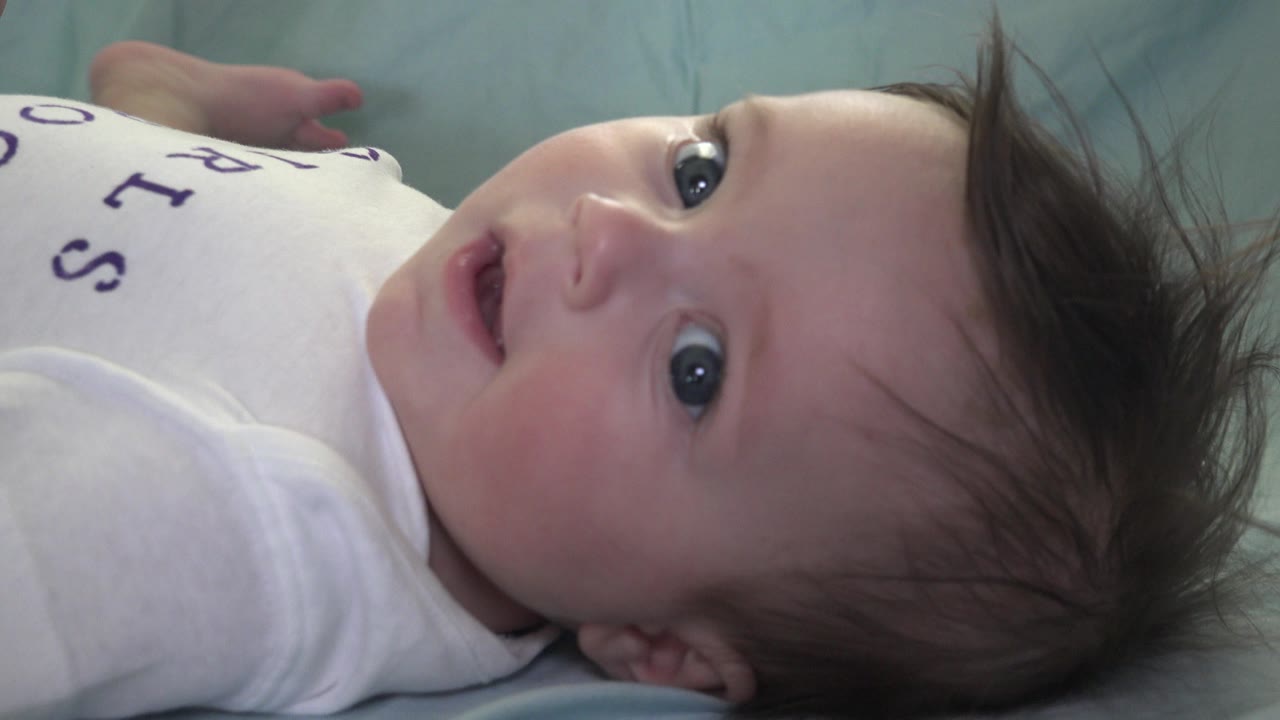 In Westminster, Colorado, a baby in a white shirt lies sideways on a light green blanket with wide eyes looking toward the camera at the start of the clip.