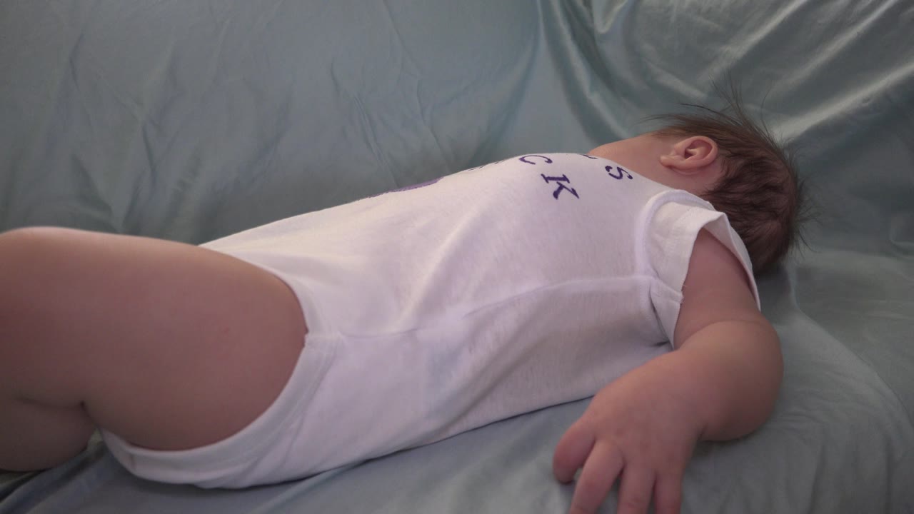 At 0:01, the baby remains lying on the pale sheet, with the white romper centered and one small arm resting closer to the camera.