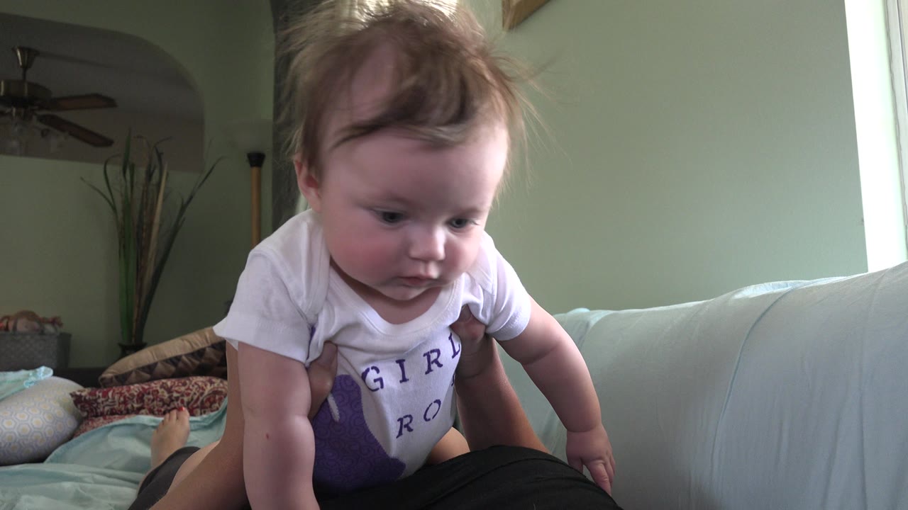 At 0:01 in Westminster, Colorado, the baby looks down while braced over the adult's black-clad lap, with the pale couch and window light remaining in view.