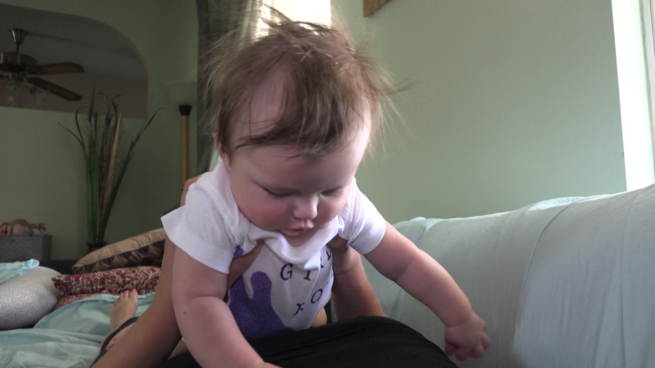 At 0:01, the baby looks down toward the parent's lap while adult hands support them on the pale couch in the Westminster, Colorado living room or bedroom setting.