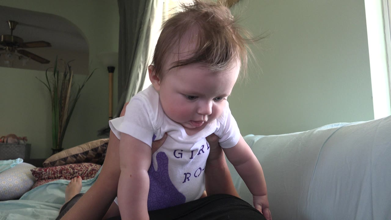 A baby in a white shirt is held above a sofa in a Westminster, Colorado living room, with green walls, curtains, and window light behind them.