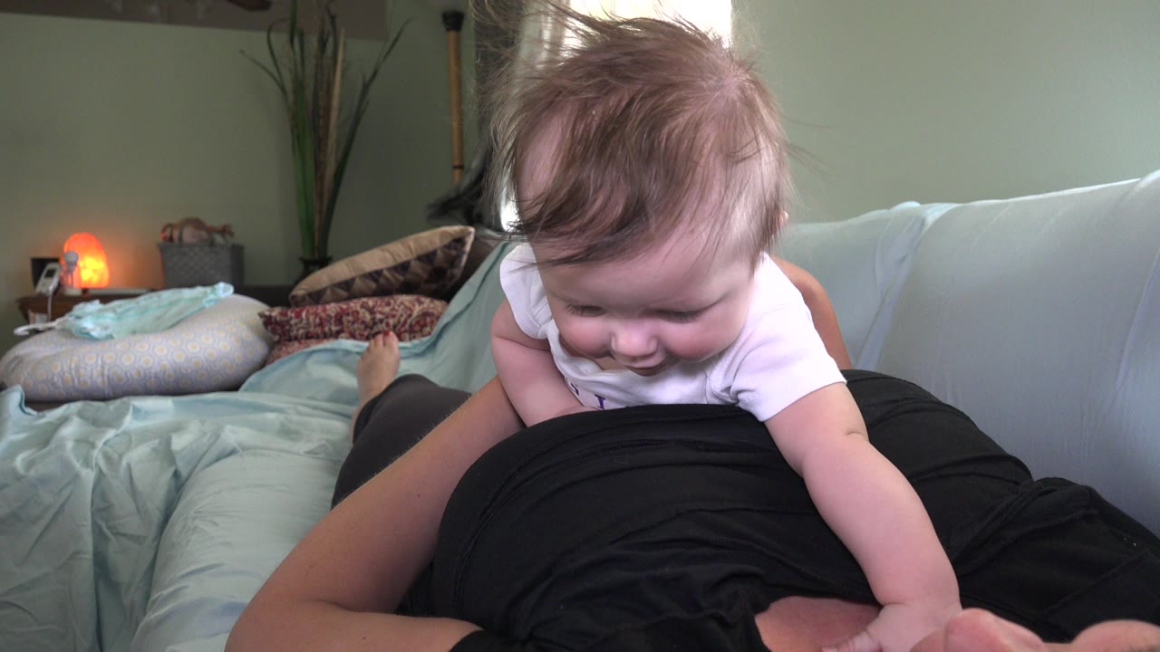 In Westminster, Colorado, a baby in a white onesie leans forward over an adult in a black shirt on a bed with pillows and a warm lamp behind them.