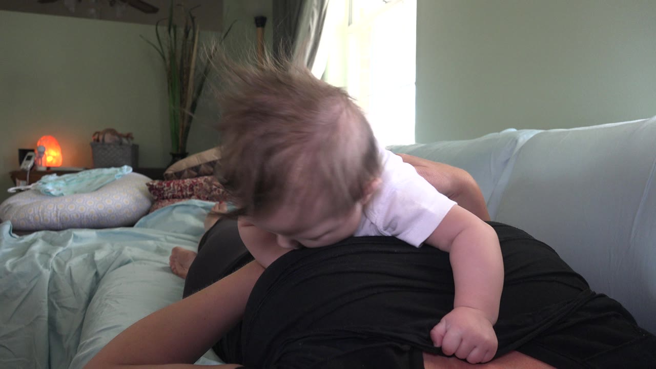 Baby leaning forward on an adult's lap in a softly lit room.