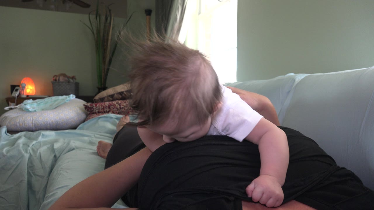 At 0:01 in Westminster, Colorado, the baby remains bent forward, one arm draped across the adult's lap, with pillows, window light, and the glowing lamp behind.