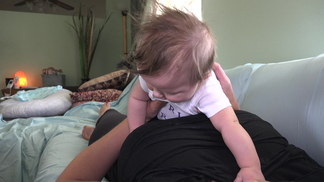 At 0:01 in Westminster, Colorado, the baby looks down toward the adult's lap while one small arm reaches outward and the bedroom pillows, wall, plant, and lamp remain visible behind them.