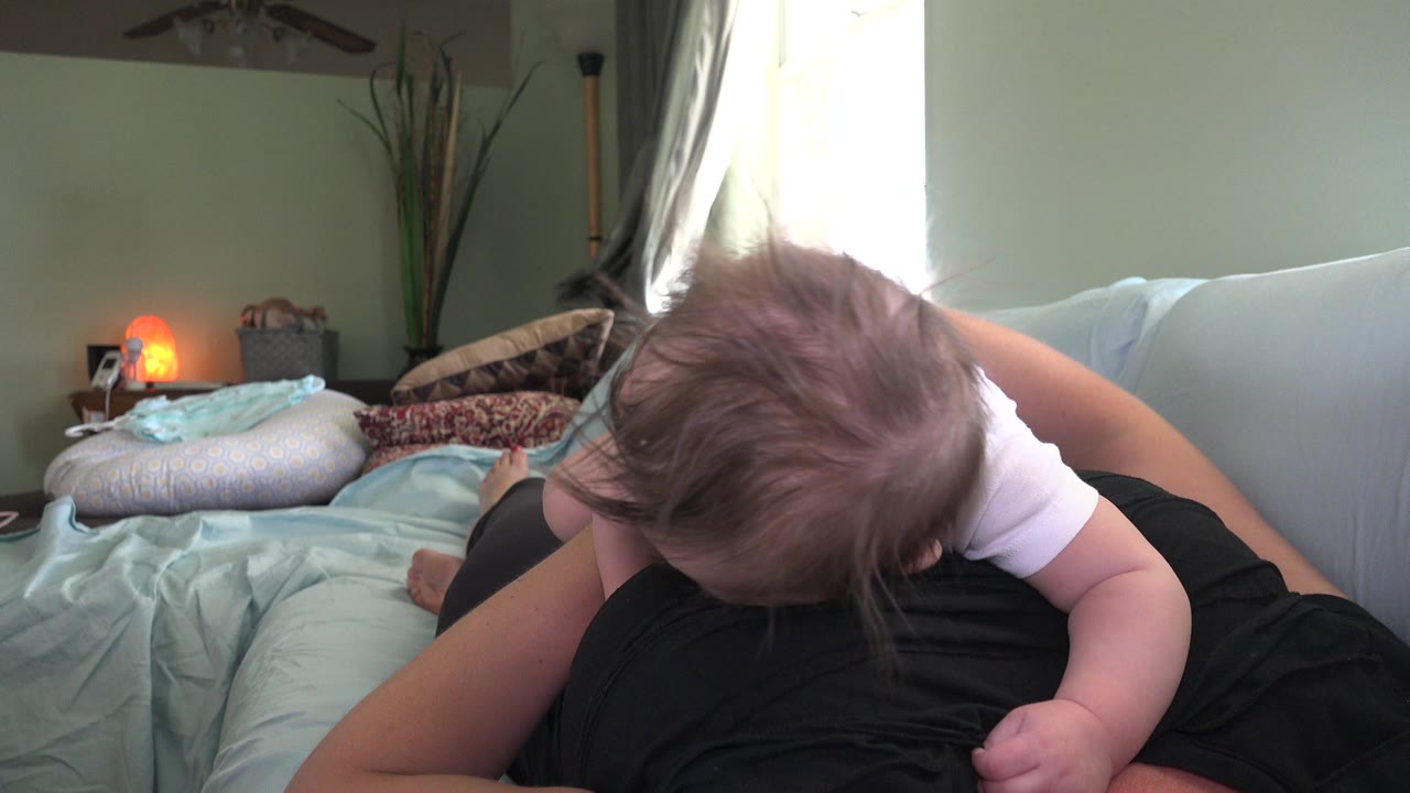 Baby leaning over an adult on a couch in a softly lit room.
