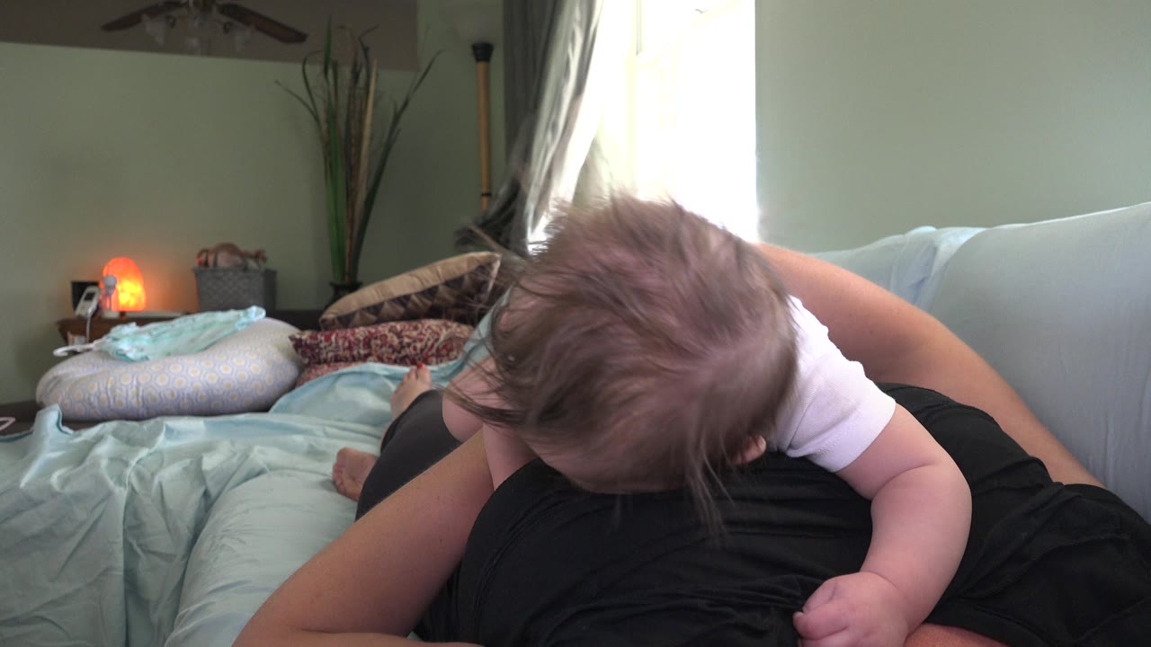 At 0:01 in Westminster, Colorado, the baby remains bent forward over the adult's torso, with the light couch, patterned pillows, plant, and bright window still visible.