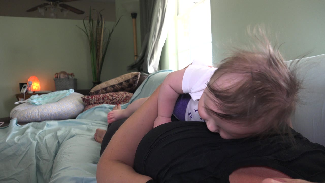Baby in white leaning over a woman in a black shirt on a bed.