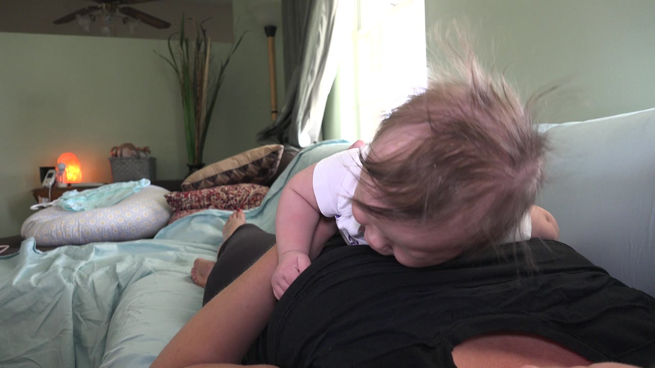 Infant in a white shirt lying on an adult's stomach on a bed.