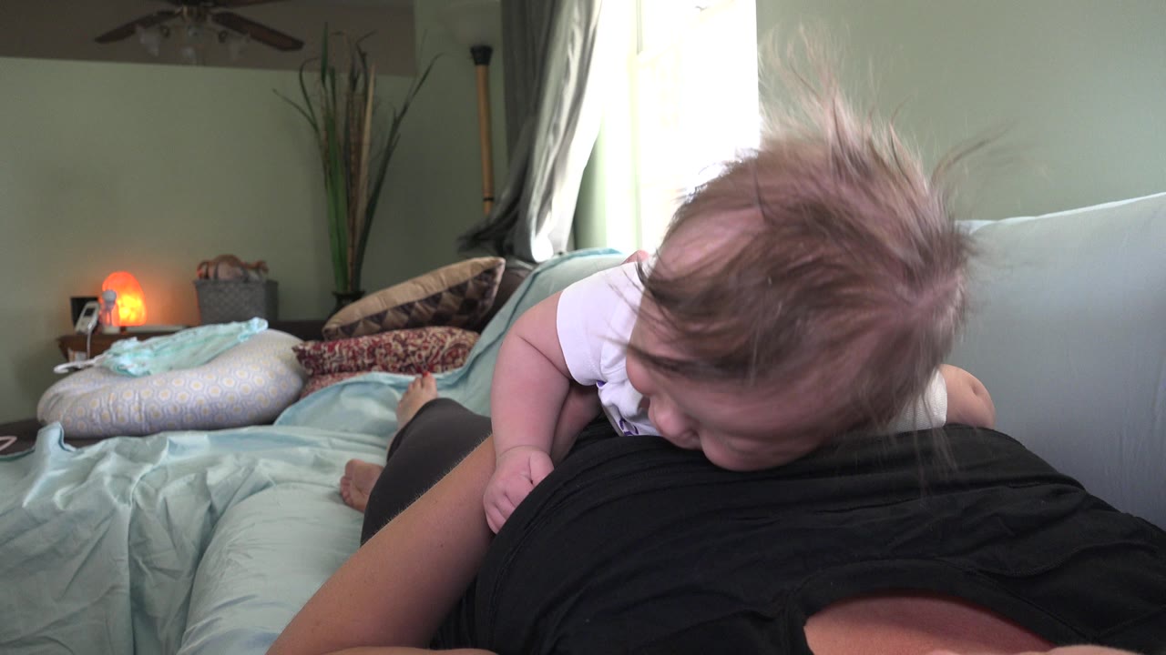 At 0:01 in Westminster, Colorado, the infant stays low against the adult's black shirt while the bed, pillows, window light, and small orange lamp remain visible in the room.