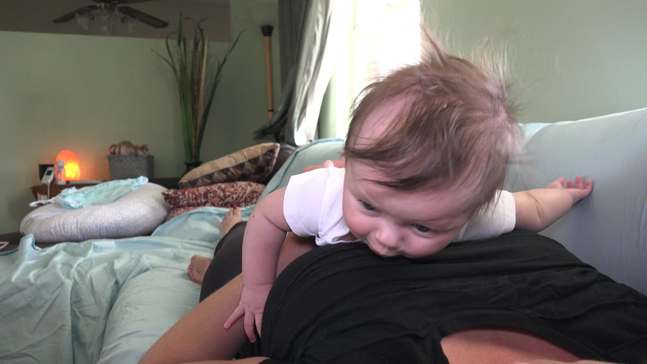 At the start of the Westminster, Colorado clip, a baby in a white onesie lies across an adult in a black shirt on a pale blue sofa with pillows and a warm lamp in the background.