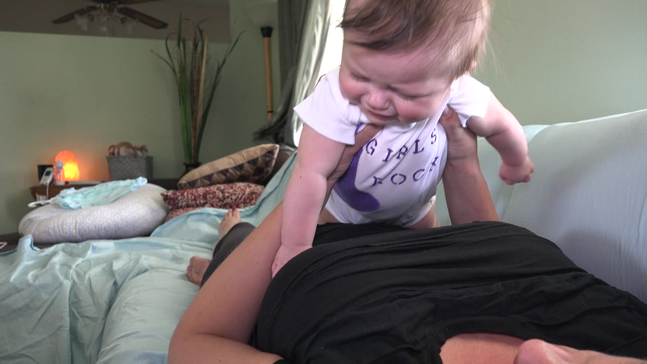 A baby in a white shirt leans over an adult lying on a sofa inside a Westminster, Colorado home, with pillows, a lamp, and a green wall behind them.