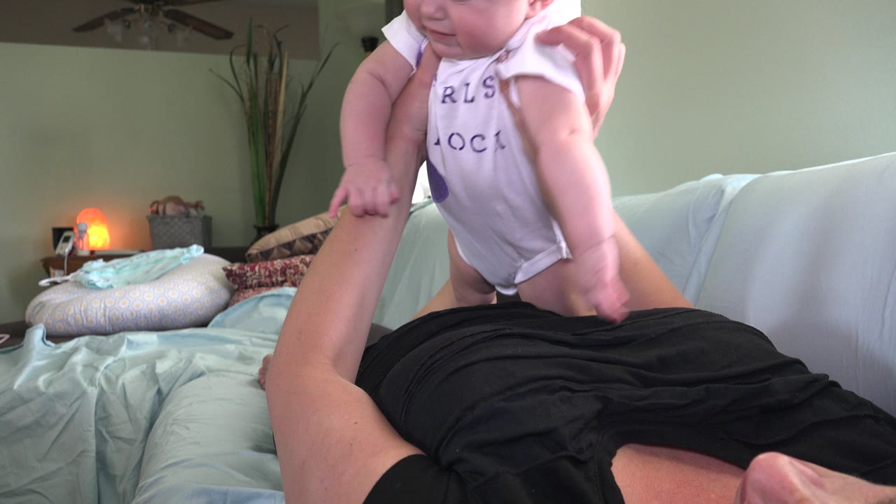 An adult's hands lift the baby slightly above the sofa, showing the baby's white shirt and legs against the pale sofa and softly lit room.