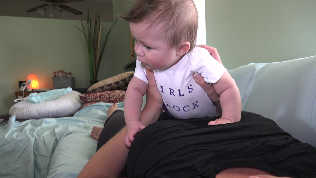 At 0:00 in Westminster, Colorado, a baby in a white shirt is held over an adult lying on a couch, with pillows, bedding, a green wall, and a glowing lamp behind them.