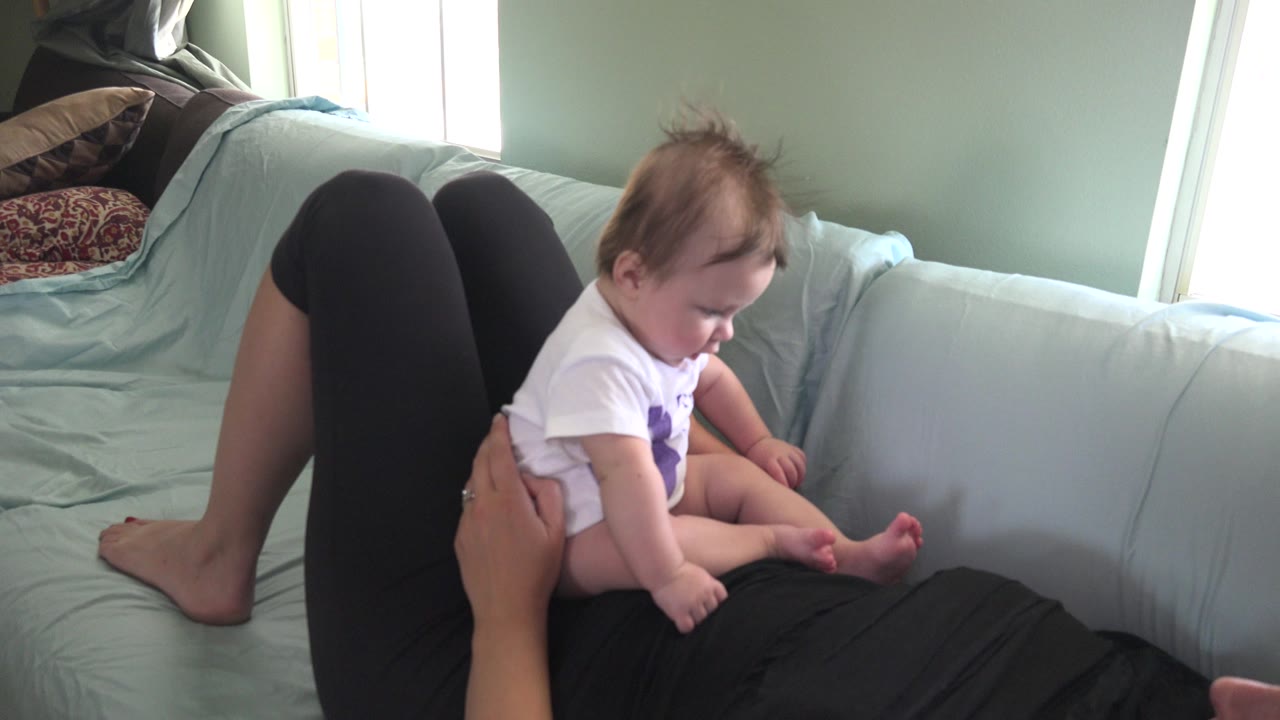One second into the Westminster, Colorado footage, the baby looks down toward their feet while an adult hand steadies them on the pale green sofa.