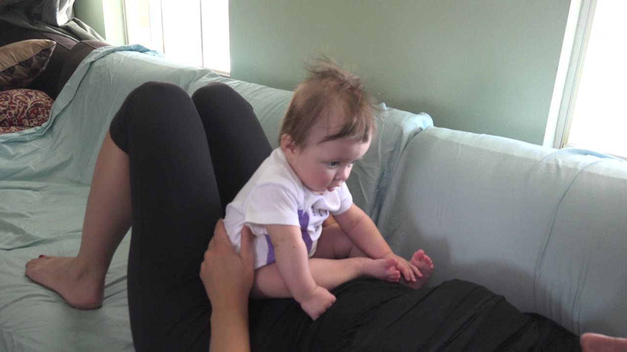 Baby sitting on an adult lying on a pale couch.