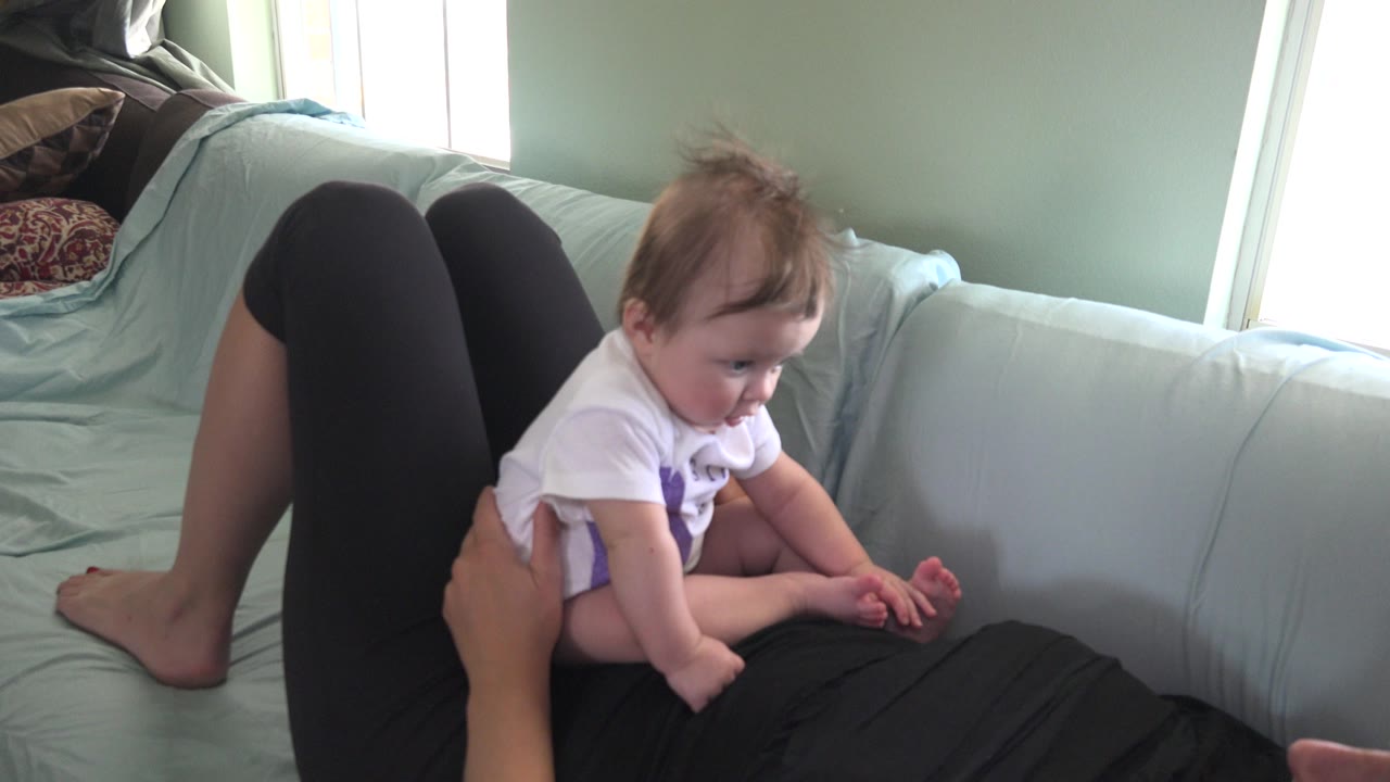 At 0:01 in Westminster, Colorado, the baby leans forward slightly on the adult's torso, with bent legs, pale couch cushions, and bright windows behind.