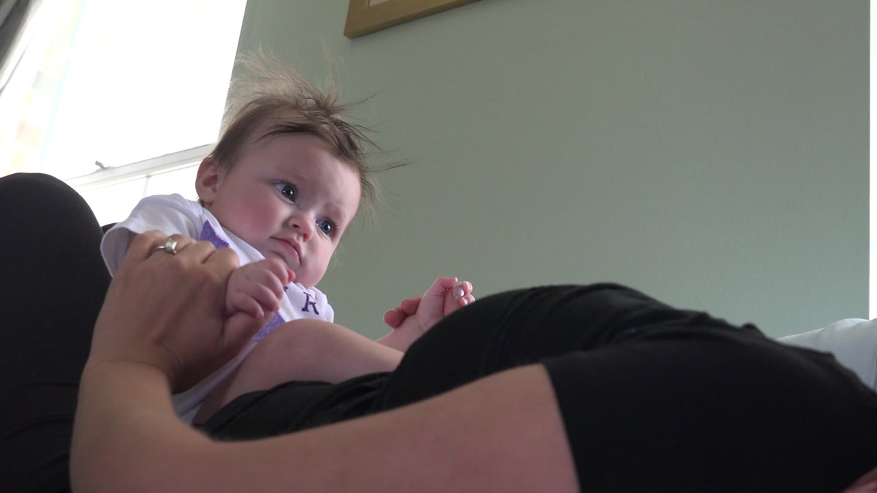 At 0:00 in Westminster, Colorado, a baby in a white shirt looks toward the camera while an adult holds the child from a low living-room angle.
