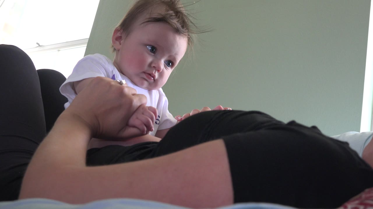 Baby lying on a woman's stomach while holding her hand indoors.