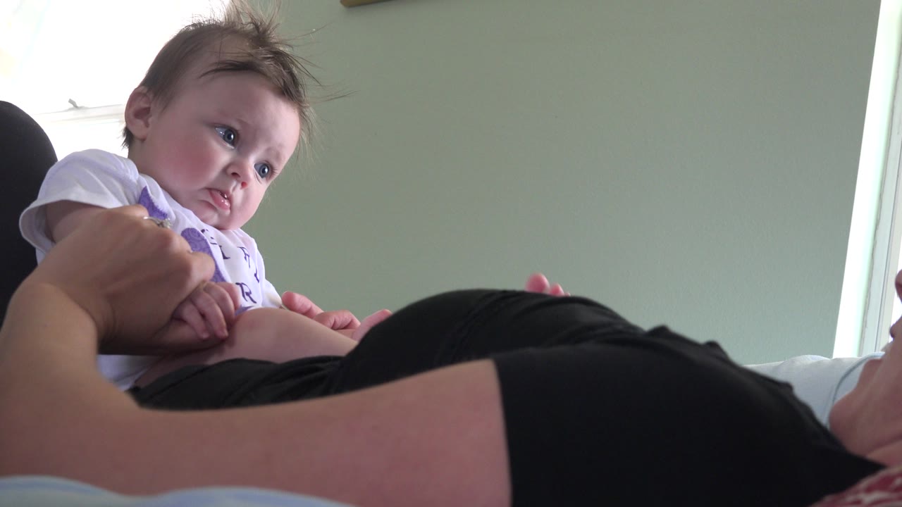 Baby leaning over a reclining woman on a bed.