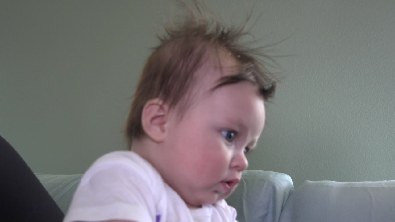 Baby in a white top leaning forward beside a pale couch.