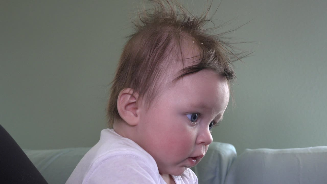 Baby with messy hair looking down while seated indoors on a couch.