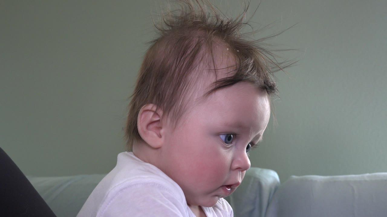 At 0:01, the baby remains in close profile on the couch, with lifted strands of hair and a focused sideways gaze in the Westminster, Colorado home interior.
