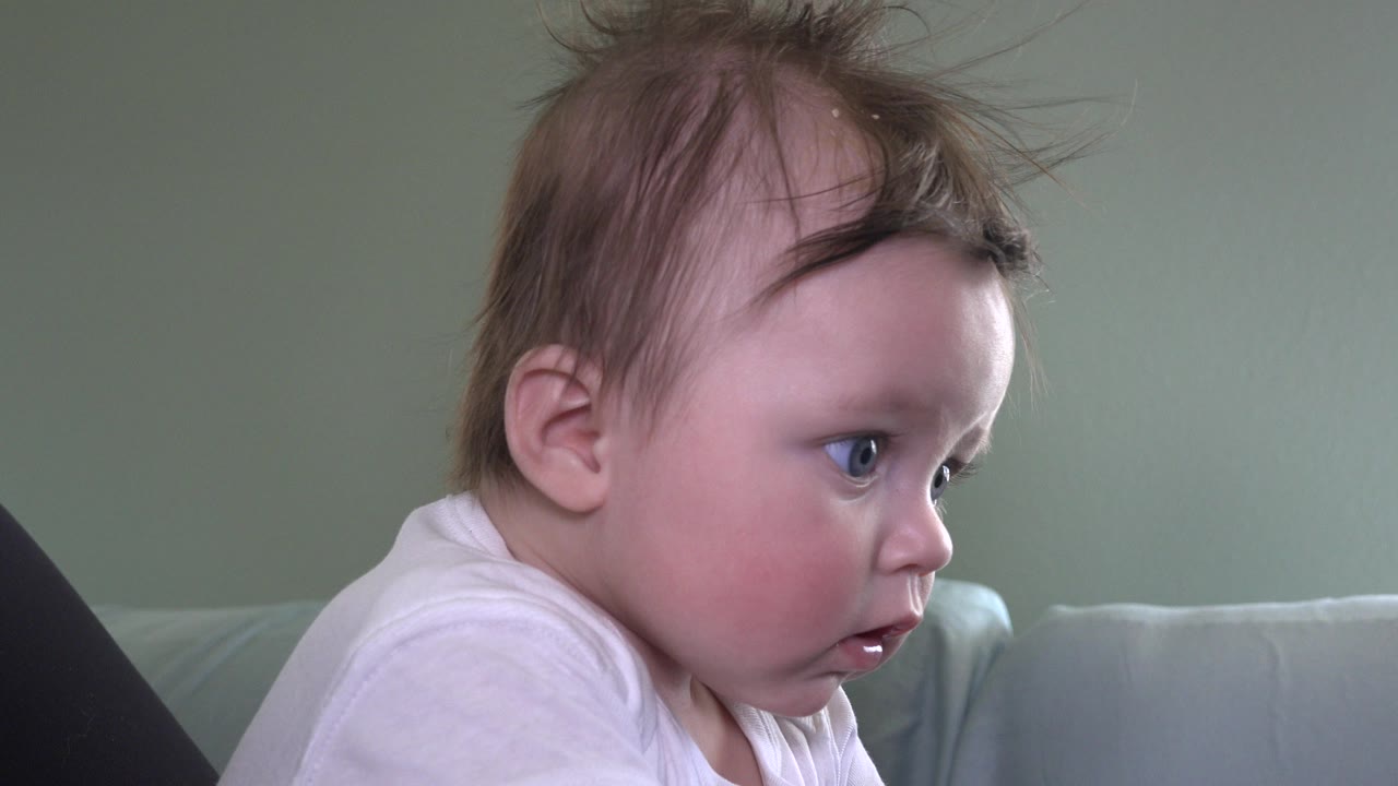 The baby remains in side profile on the couch in Westminster, Colorado, with wide eyes and tousled hair visible under soft indoor light.