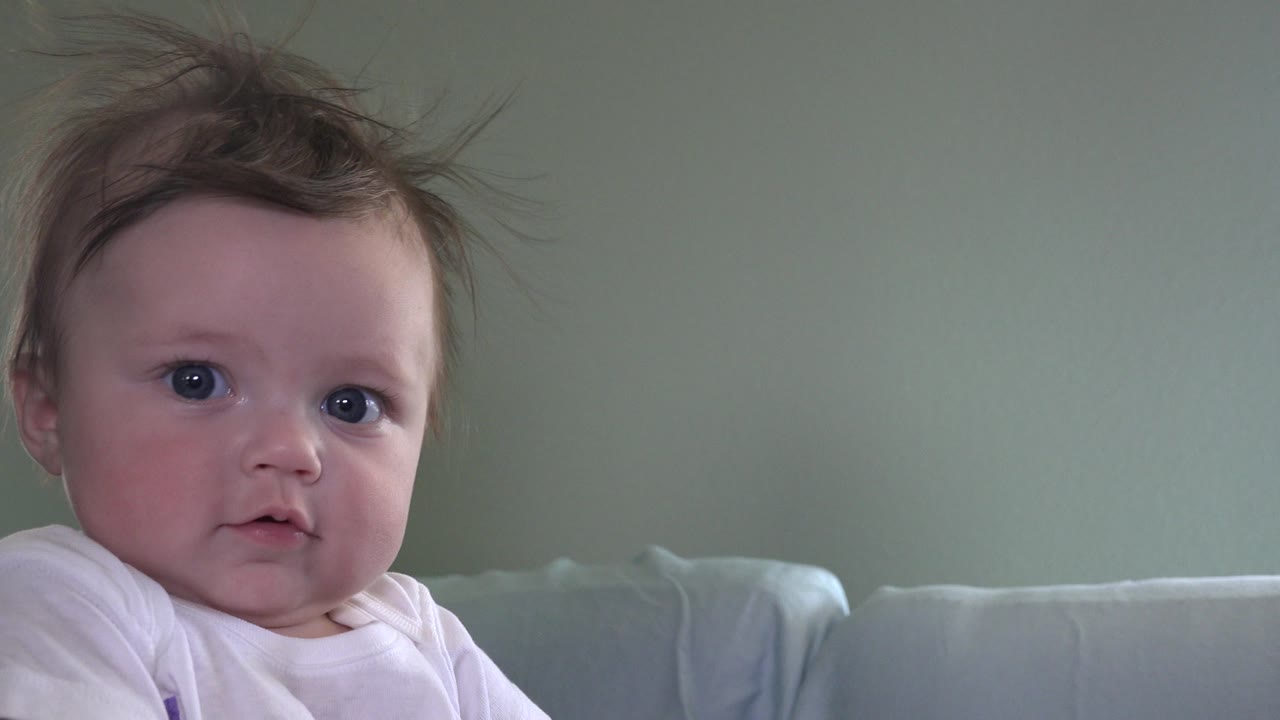Baby in a white shirt looking toward the camera indoors.